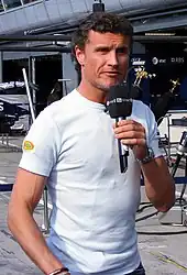 David Coulthard holding a microphone in his left hand speaking to the British media