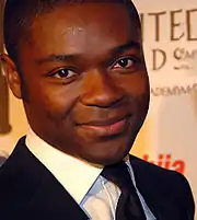 A short dark-hair black man wearing a suit with black jacket and tie, over a white shirt, is looking straight to the camera smiling.