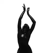 A black-and-white photo of Richard covered in dark black body paint, nude, and posing with her arms raised to the air