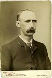 A man in a suit and tie is shown from the chest up, facing slightly right of the camera.