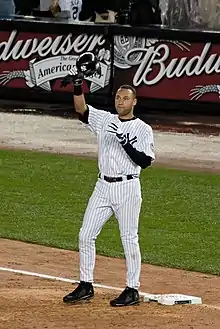 A man in a white baseball uniform with navy pinstripes removes his helmet to salute the crowd, which is cheering for him.