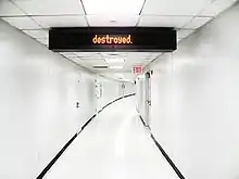 A white hallway, with doors and a red EXIT sign on the right side. On the ceiling, a sign with orange LED text reading "destroyed."