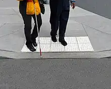 Two people stand on top of light-colored truncated domes at a curb cut.  They both face the parking lot, and the person in front holds a white cane.