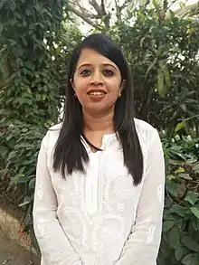 This is a waist length photograph of a brown woman with long hair. Her hands are placed close to her body. She is wearing a white kurta and red lipstick. She is smiling at the camera. There are trees in the background.