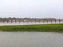 A reddish metal bridge supported by a row of narrow columns crosses horizontally over a stretch of river water, with a grass-covered mudbar in the foreground