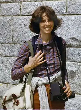 A photo of Diana Mara Henry standing with camera equipment draped over her shoulders.