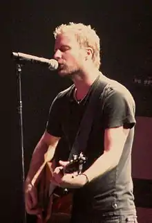 A fair-haired man singing into a microphone and playing a guitar