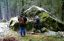 Two people with their backs to the viewer stand in front of a large boulder in the middle of woodland. One of the figures is pouring a liquid onto the ground.