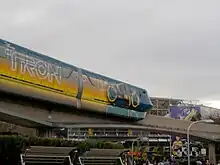 A monorail stamping a painted light cycle which leaves an orange trail behind. The second cart also has the Tron: Legacy title.