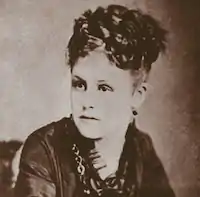 an old photograph of a woman in 3/4 view tightly cropped to just her face, she is in a dark dress with curly hair pinned on top of her head.