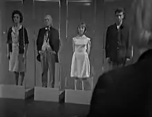 Copies of Barbara, the Doctor, Vicki, and Ian stand upright, emotionless, inside glass boxes. The Doctor's shoulder can be seen overlooking them.