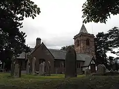 A church seen from the north; to the right is a tower with a small pyramidal spire; projecting from the church are a gabled transept and a timber-framed porch. In the foreground are gravestones
