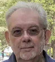 color photo of an older white man with glasses and a beard looking into the camera