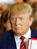 Official portrait of Donald Trump wearing a navy blue suit and smiling in front of the United States flag.