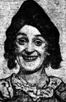 A grinning woman wearing white makeup and a dark curled wig