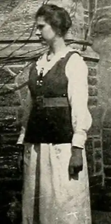 A young white woman with dark hair, standing outdoors in front of a brick wall, wearing a dark belted vest over a white blouse and skirt