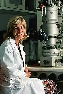 A smiling blonde older woman in a lab coat sits at an electron microscope.