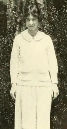 A young white woman, standing, hair center-parted; wearing a white top and skirt, with a round collar and long sleeves