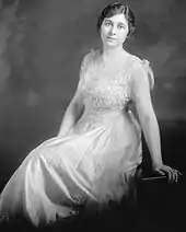 A slightly built, dark haired woman wearing a white dress and leaning backward onto her left arm