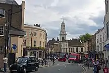 A curving street with older two- and three-storey buildings on either side. In front is a black London taxicab with an advert; midway down the street is an intersection with heavy traffic. A cupolaed clock tower rises in the rear