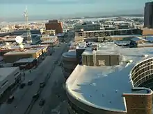 Snow-covered flat roofs and snowy streets seen from slightly above. In the far background is a frozen lake and some low hills