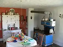 Interior of kitchen: cupboard against wall, table and chairs in foreground, cast-iron stove with pipe leading to chimney