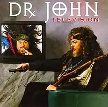 Dr. John sitting in an easy chair, holding a remote, and watching himself on television