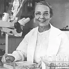An older white woman, smiling, at a microscope.