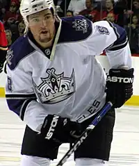 Hockey player in white and dark blue uniform. He crouches to his right, holding his stick across him.