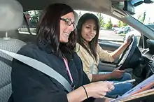 Two women sit in a car.  The passenger holds a paper so the driver can see it.