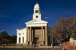 Modified cruciform plan form church with hipped roof. Belfry set in roof.
Symmetrical facade with stone portico.
Rough coarse stone base. Plastered core with pilasters and cornice. Plastered area is ruled and joined. Pediments above door openings
Tall arc The foundation stone was laid on 29 November 1830 by W C Rhyneveldt, the Civil Commissioner of Graaf
Type of site: Church
Current use: Religious.
The church is the major landmark in Colesberg and is closely associated with the development of the[clarification needed]