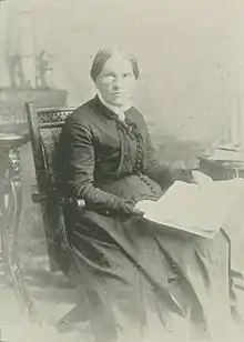 An older white woman, seated, holding a large book open on her lap.
