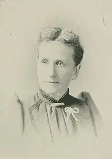 B&W portrait photograph of a middle-aged woman with hair in an up-do wearing a high-collared blouse with a white ribbon pinned on the left side.