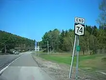 An intersection of a pair of highways in a wooded rural area. In the foreground is a sign assembly reading "End NY 74"; in the foreground is a second assembly indicating that Interstate 87 is straight ahead and US 9 is accessed by turning either left or right.