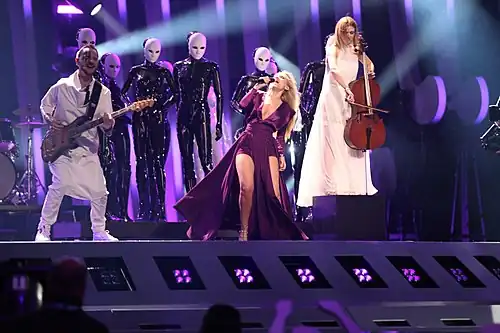 Two women—dressed in purple and white dresses, respectively—performing on a stage alongside a guitarist; black mannequins are seen in the background.