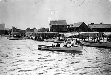 family in a row boat in front of a coastline filled with docks, nets, and fish houses