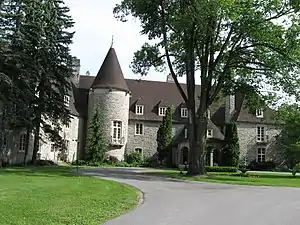 In the foreground is a mowed lawn to the left and a paved road to the right. The road branches to circle an island of grass containing a large tree and a small fountain. A three-storey grey stone building with a brown roof is prominent behind them. Its entrance is partially obscured by the tree, and a three-storey rotunda is clearly visible fronting the right side of the building.