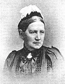 Photograph of a woman wearing a close fitting feathered cap and a dark dress clasped at the neck with a broach.