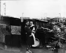 Black and white photograph of a woman wearing traditional Japanese clothing holding a small child while standing in front of a crudely built shack. Rubble and undamaged houses are visible in the background.