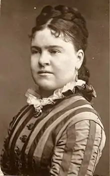 A round-faced white woman with her dark hair in an elaborate braided and curled coiffure. She is wearing a gown with prominent vertical stripes on sleeves and bodice.