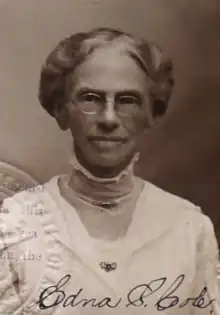 An older white woman with grey hair dressed to the nape, wearing eyeglasses and a high-collared white dress