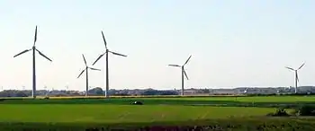 A car drives past 4 wind turbines in a field, with more on the horizon
