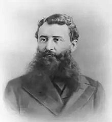 Black and white portrait of a man wearing a coat, shown from chest up, well combed and with a voluminous beard.