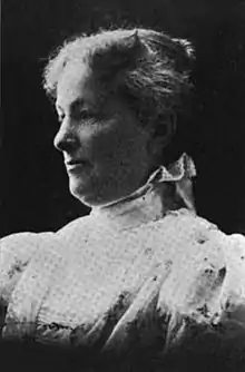 A white woman, in 3/4 profile, wearing a high-necked white dress with large sleeves.