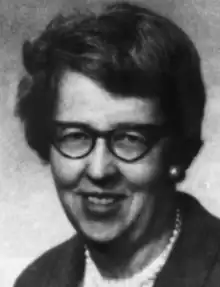 A smiling middle-aged white woman, hair in a short bouffant style, wearing glasses and pearls