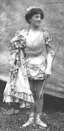 A young white woman, standing in a short corseted costume, with her hair in a bouffant updo. She is holding a stick in one hand and a cloak in the other.