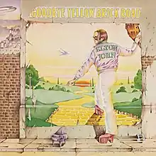 A wall with a worn poster showing a yellow brick road winding down towards the green plains and the sunset. Elton John, shown with a purple jacket bearing his name, has his right foot on the yellow brick road and his right hand holding on to the poster. On the sidewalk, a minature piano with a musical note. The album's title is placed above the poster.