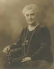An older white woman with white hair, seated in a carved wooden chair, wearing a loose-fitting dark velvet dress with long sleeves
