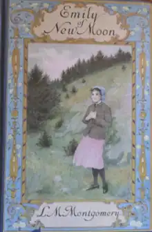 A rectangular image used for the book cover of the novel "Emily of New Moon". At the centre of the image is Emily Starr wearing a pink dress, covered by a grey coat. She is clasping her hands over her heart. She is wearing a blue bonnet on her head and standing outdoors on the base of a small hill covered in pine trees. Surrounding this painting is a blue border with intricate floral decor and brass decoration that twist to frame the title at the top.