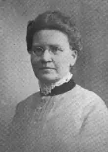 A middle-aged white woman with dark hair in a simple updo, wearing a high-collared bodice with lace trim